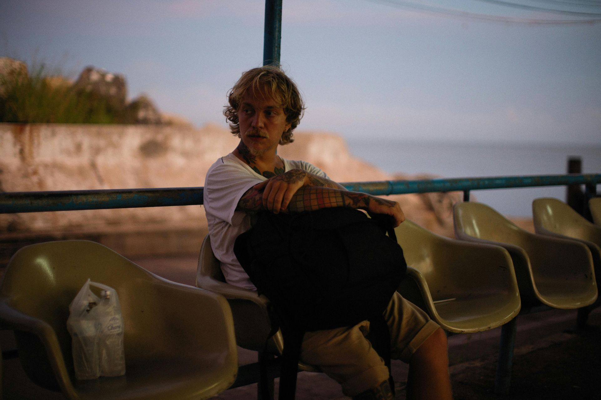 Una persona con i capelli chiari, tatuaggi e uno zaino è seduta su una sedia di plastica curva accanto a una ringhiera che si affaccia sul mare.