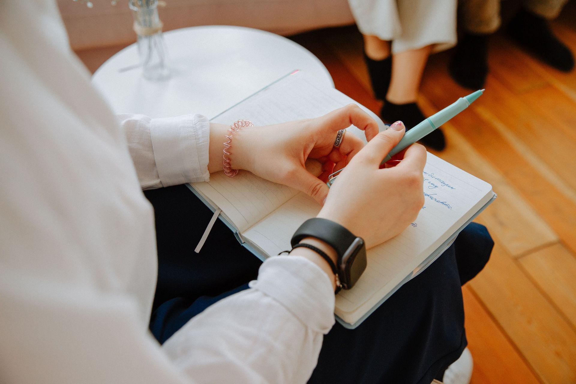 Una persona con una camicia bianca scrive su un quaderno con una penna chiara, indossando uno smartwatch e un braccialetto di perline.