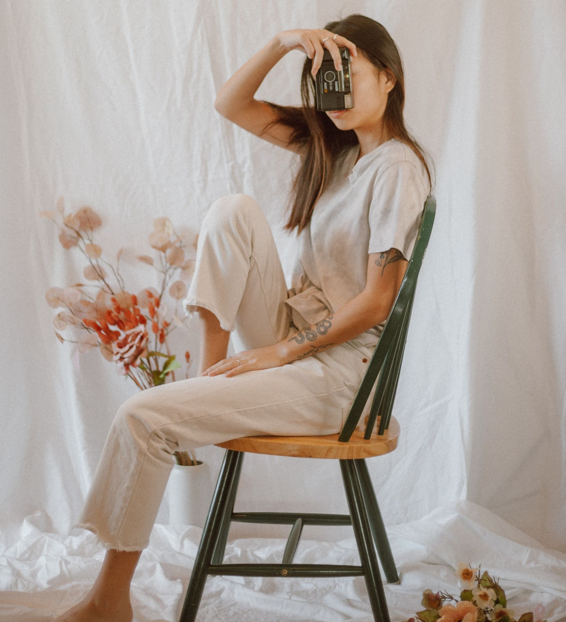 In uno studio bianco, una persona vestita di beige è seduta su una sedia a fuso verde scuro, con una macchina fotografica puntata al viso.