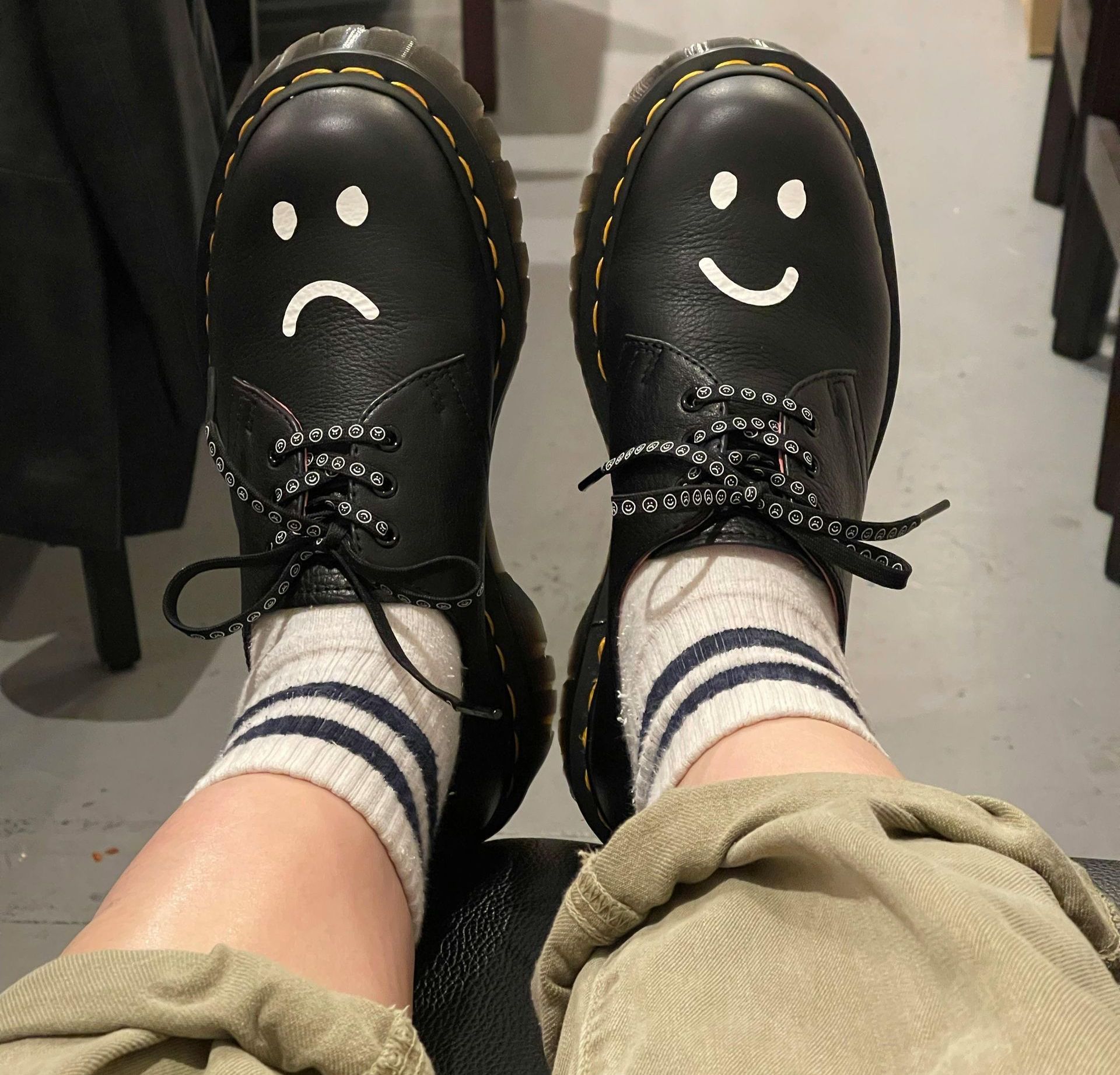 Un paio di scarpe Dr. Martens nere, ciascuna con un viso dipinto a mano; uno triste e uno felice, indossate con calzini a righe.