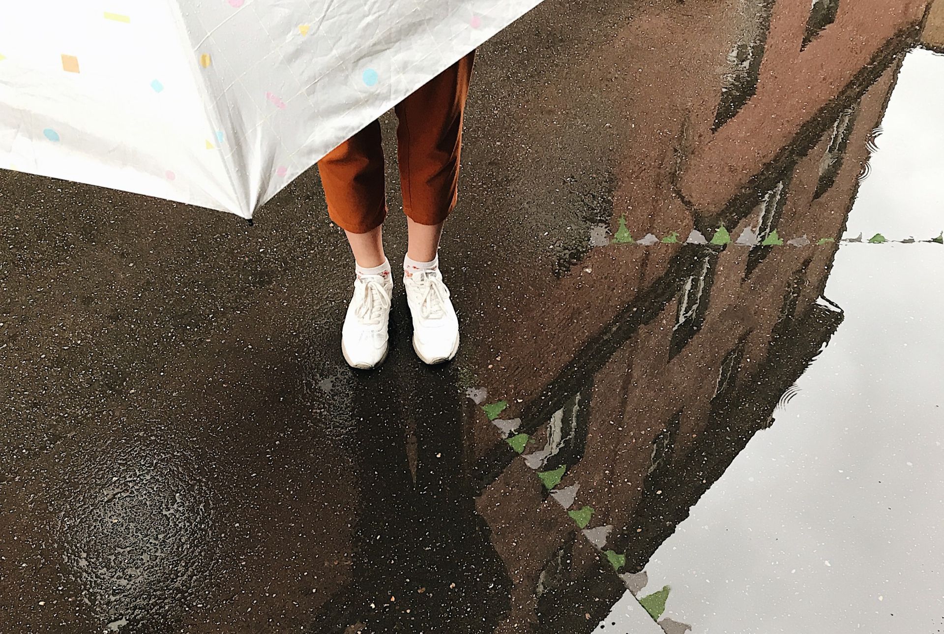 Una persona si ripara sotto un ombrello bianco su un marciapiede bagnato, il suo riflesso visibile in una grande pozzanghera.