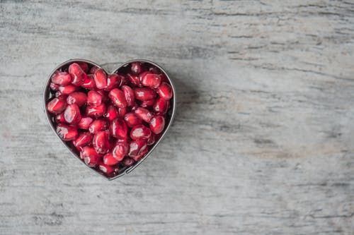 Una ciotola a forma di cuore, colma di chicchi di melograno rosso acceso, poggia su una superficie di legno grigio chiaro e dalla texture particolare.