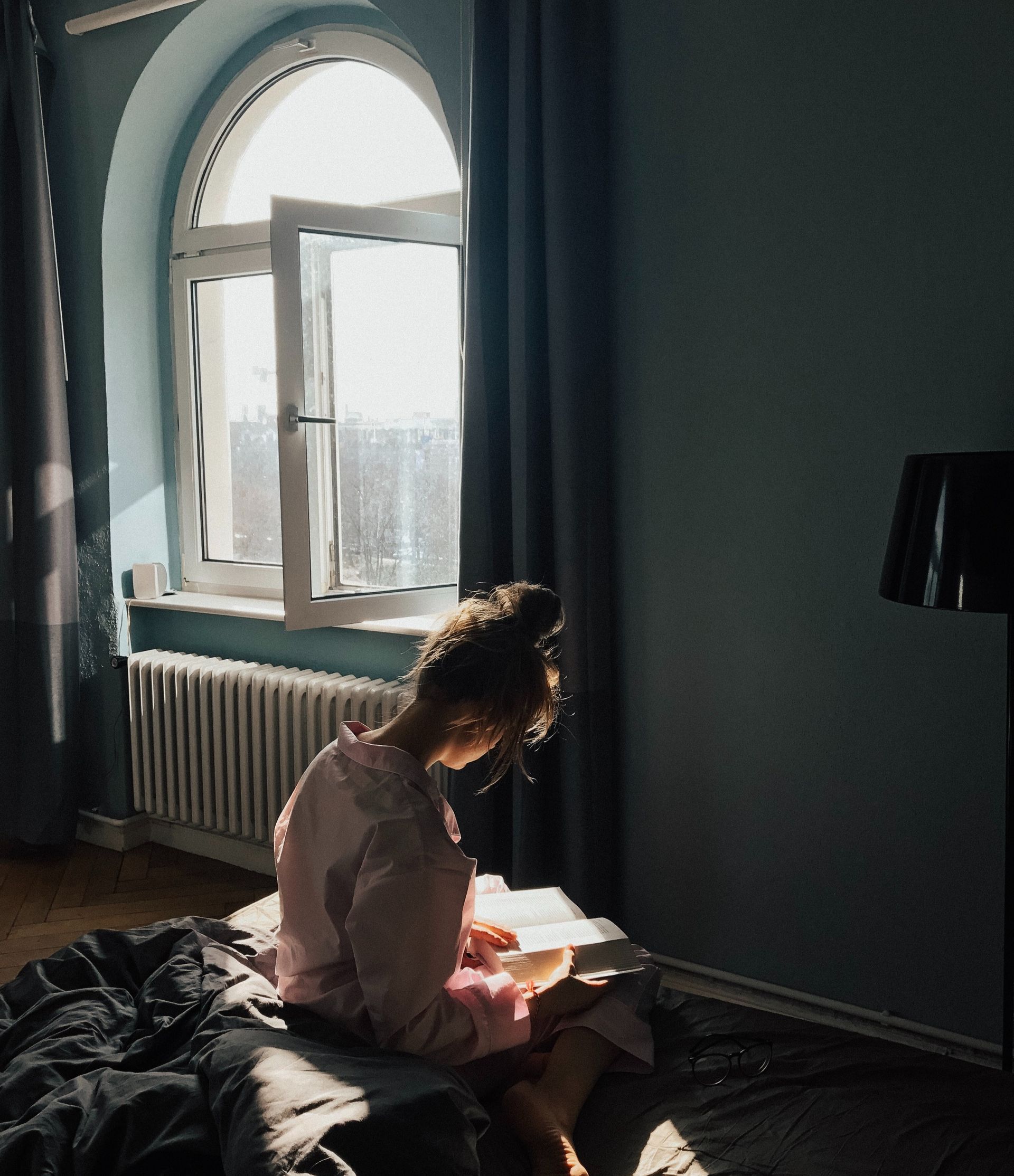 Una persona è seduta su un letto in una stanza illuminata dal sole, con le pareti blu, e legge un libro accanto a una finestra ad arco aperta.