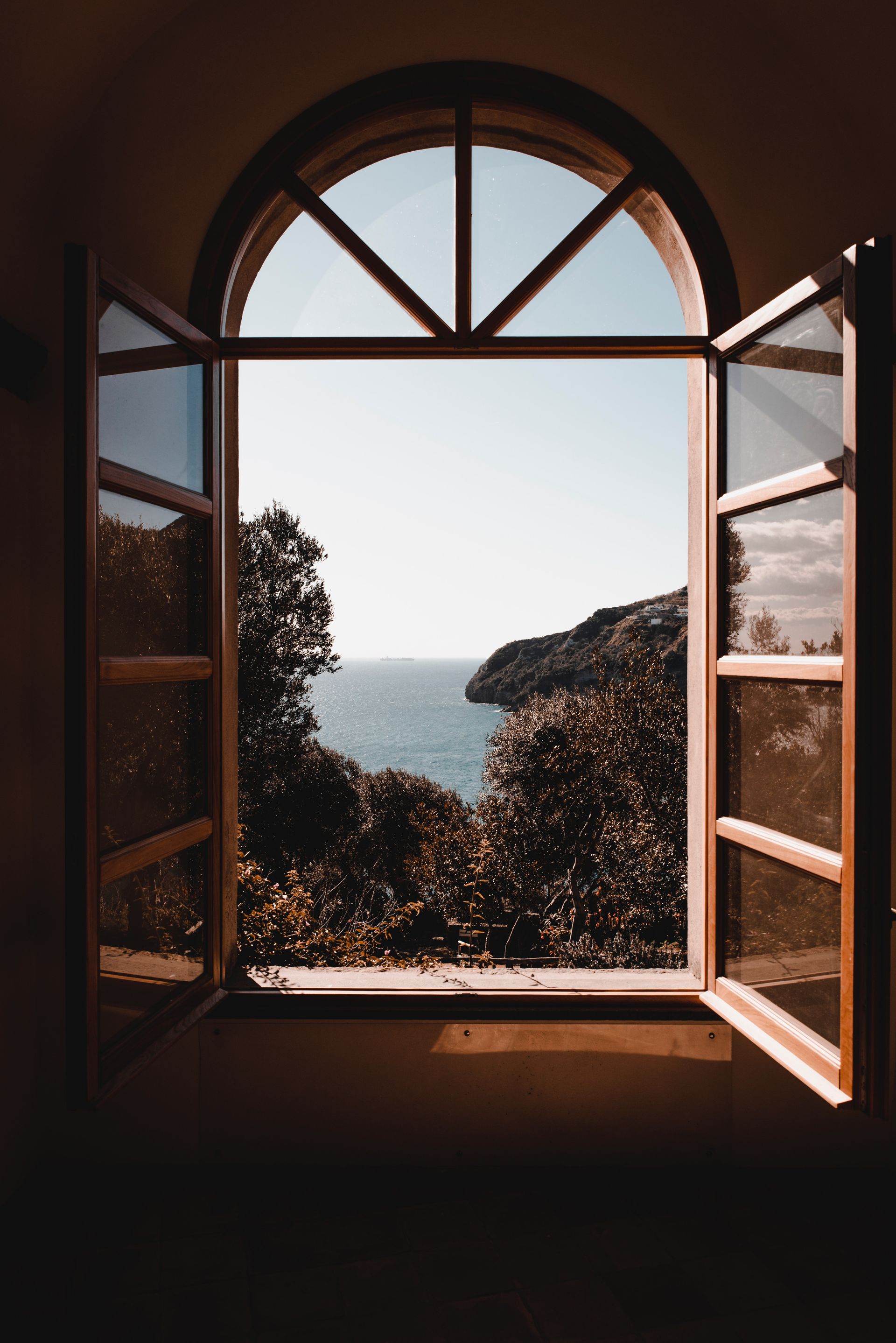 Un'ampia finestra ad arco incornicia la vista di una lussureggiante collina che si affaccia su un mare calmo, sotto un cielo limpido e luminoso.