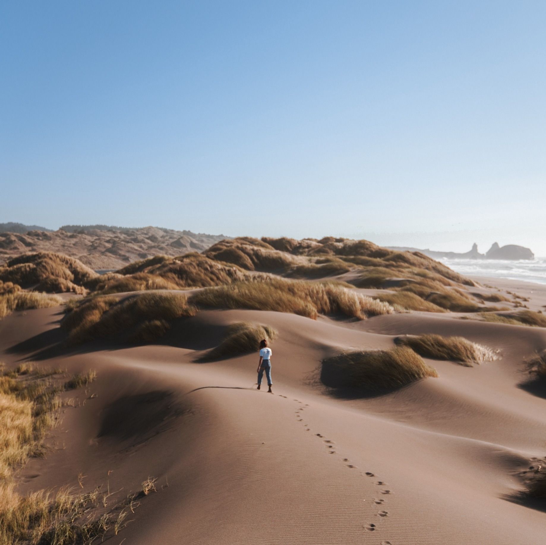 Una persona cammina lungo una scia di impronte tra dune di sabbia illuminate dal sole, con le colline costiere e l'oceano in lontananza.