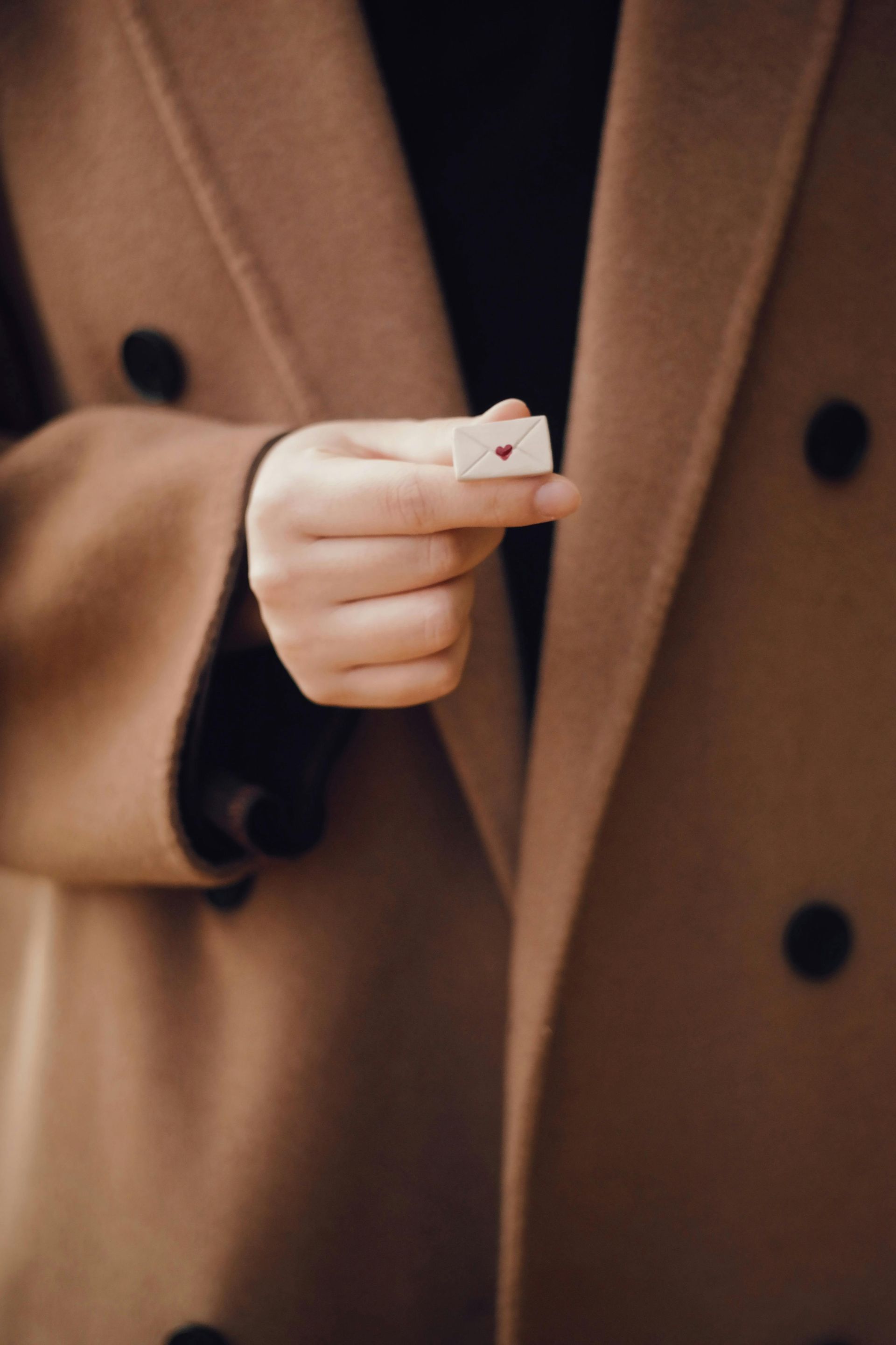Una mano avvolta in un cappotto color cachi stringe una minuscola busta beige con un piccolo cuore rosso sulla patta.