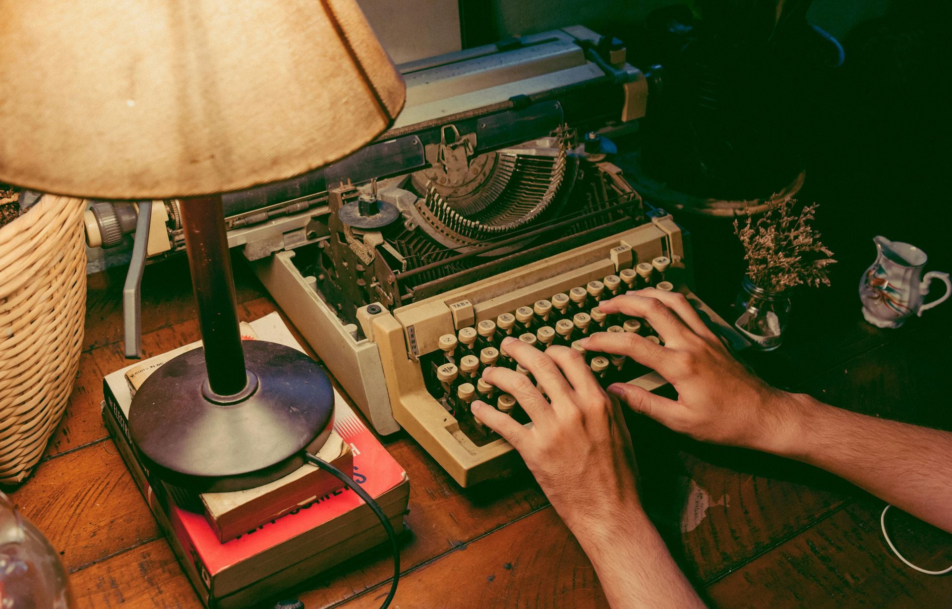 Le mani di una persona digitano su una macchina da scrivere d'epoca a una scrivania illuminata da una lampada, con libri e una piccola brocca nelle vicinanze.