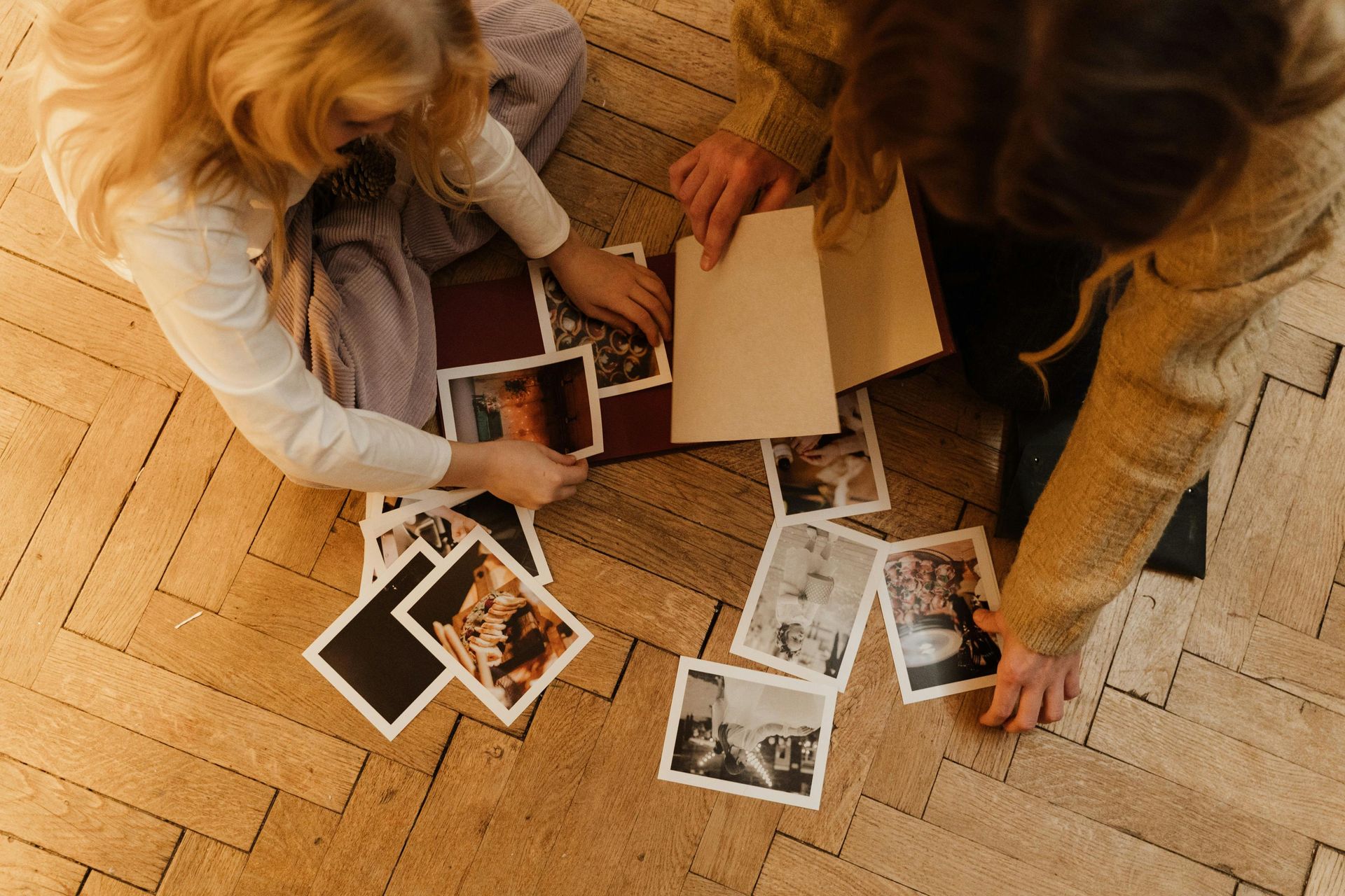 Due persone sono sedute su un pavimento di parquet, intente a riordinare una collezione di piccole fotografie e una cartella di documenti.