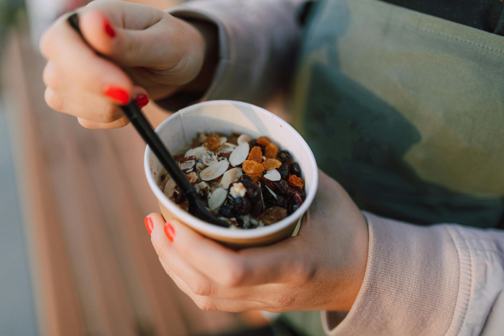 Una persona tiene in mano un bicchiere di carta contenente un mix di frutta secca con mandorle a lamelle e frutta disidratata, mentre usa un cucchiaio di plastica nero.