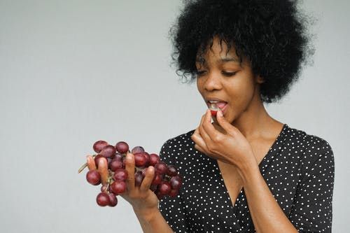 Una persona con i capelli ricci che indossa una camicia a pois tiene in mano un grappolo d'uva rossa e ne mangia uno.