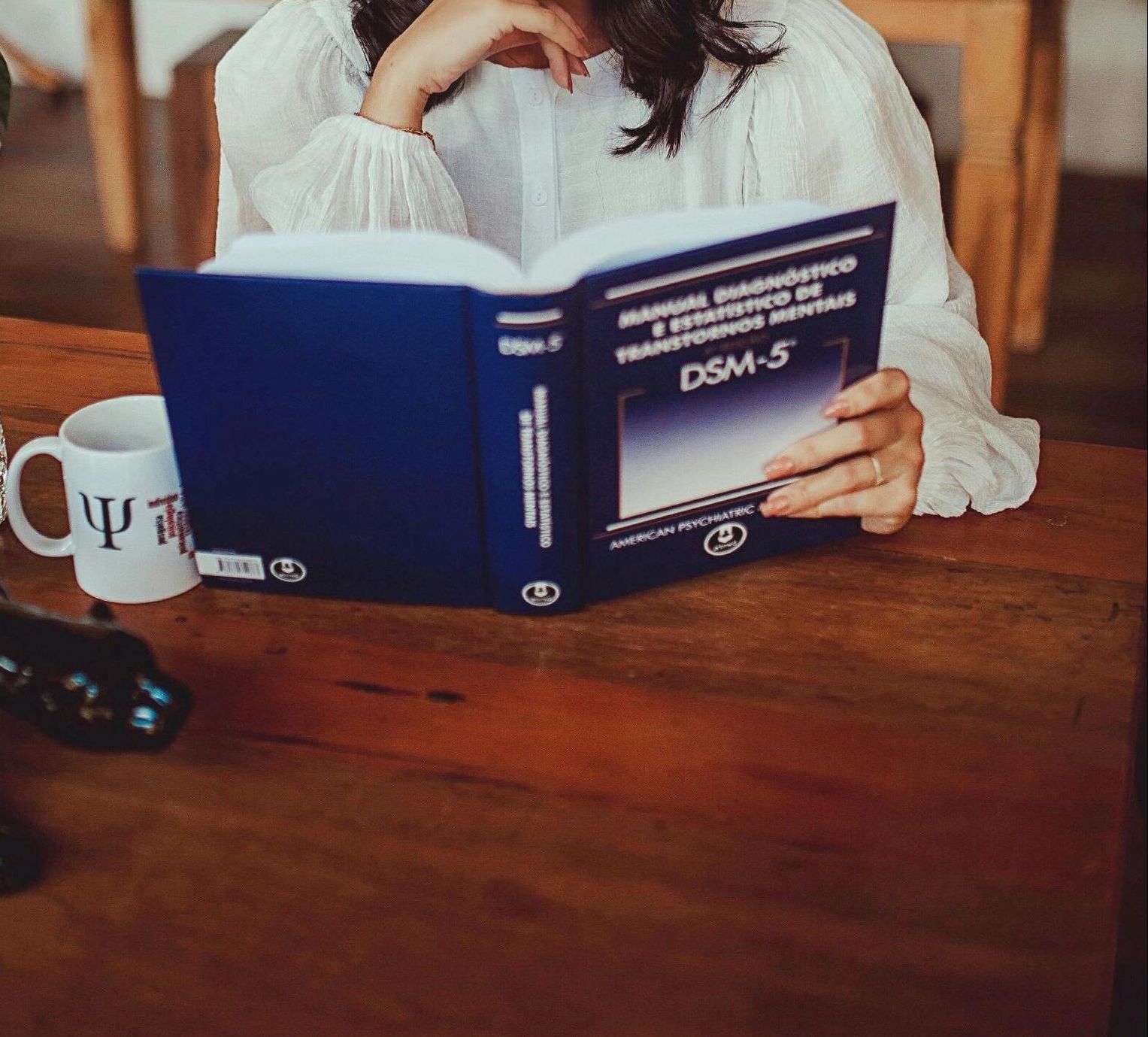 Una persona con una maglietta bianca legge un libro blu del DSM-5 seduta a un tavolo di legno con una tazza da caffè a tema psicologico.