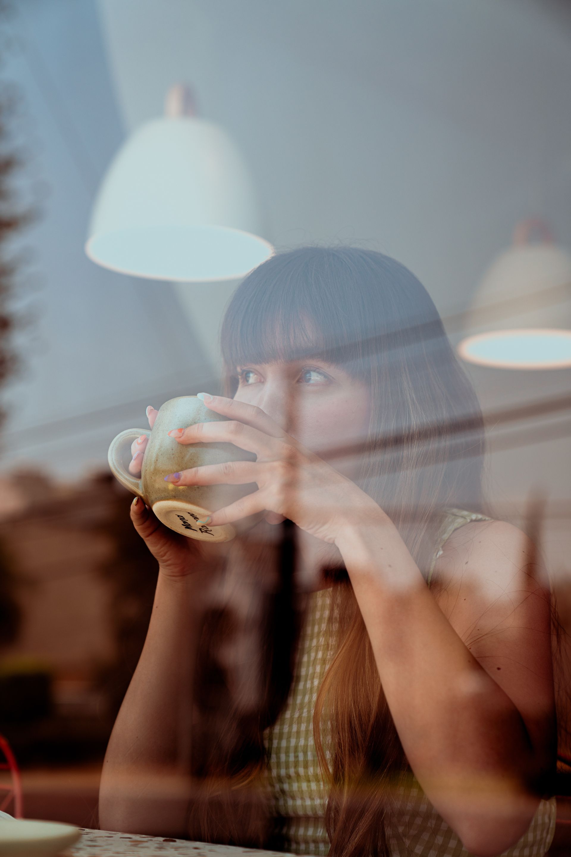 Una persona con i capelli scuri e la frangia beve da una tazza, vista attraverso il riflesso di un vetro in un ambiente accogliente e dalla luce soffusa.