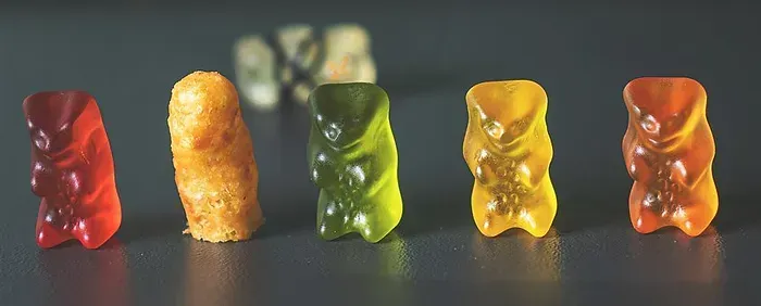 Una fila di quattro orsetti gommosi rossi, verdi, gialli e arancioni, con uno snack giallo ruvido e dalla consistenza particolare posizionato tra i primi due.