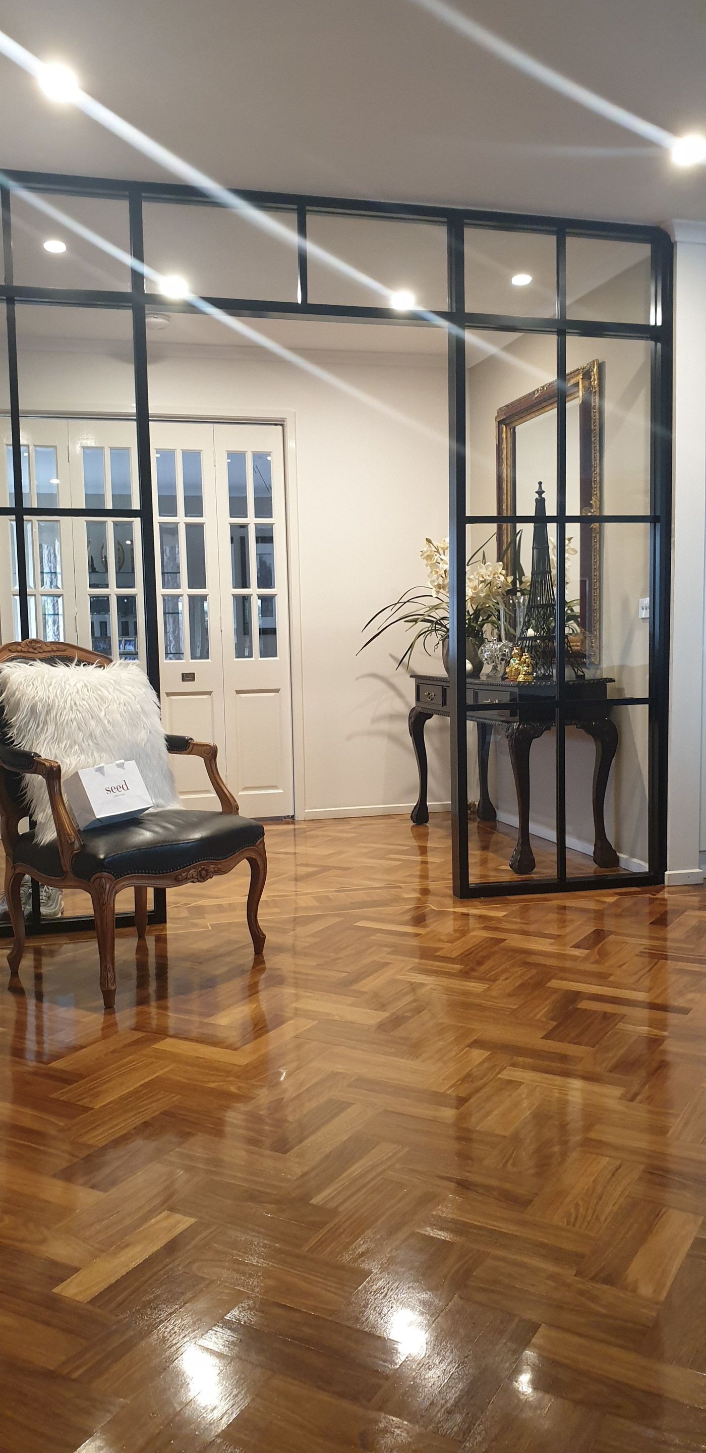 A polished parquet floor leads to an entryway framed by a black metal-grid partition, a chair, and a console table.