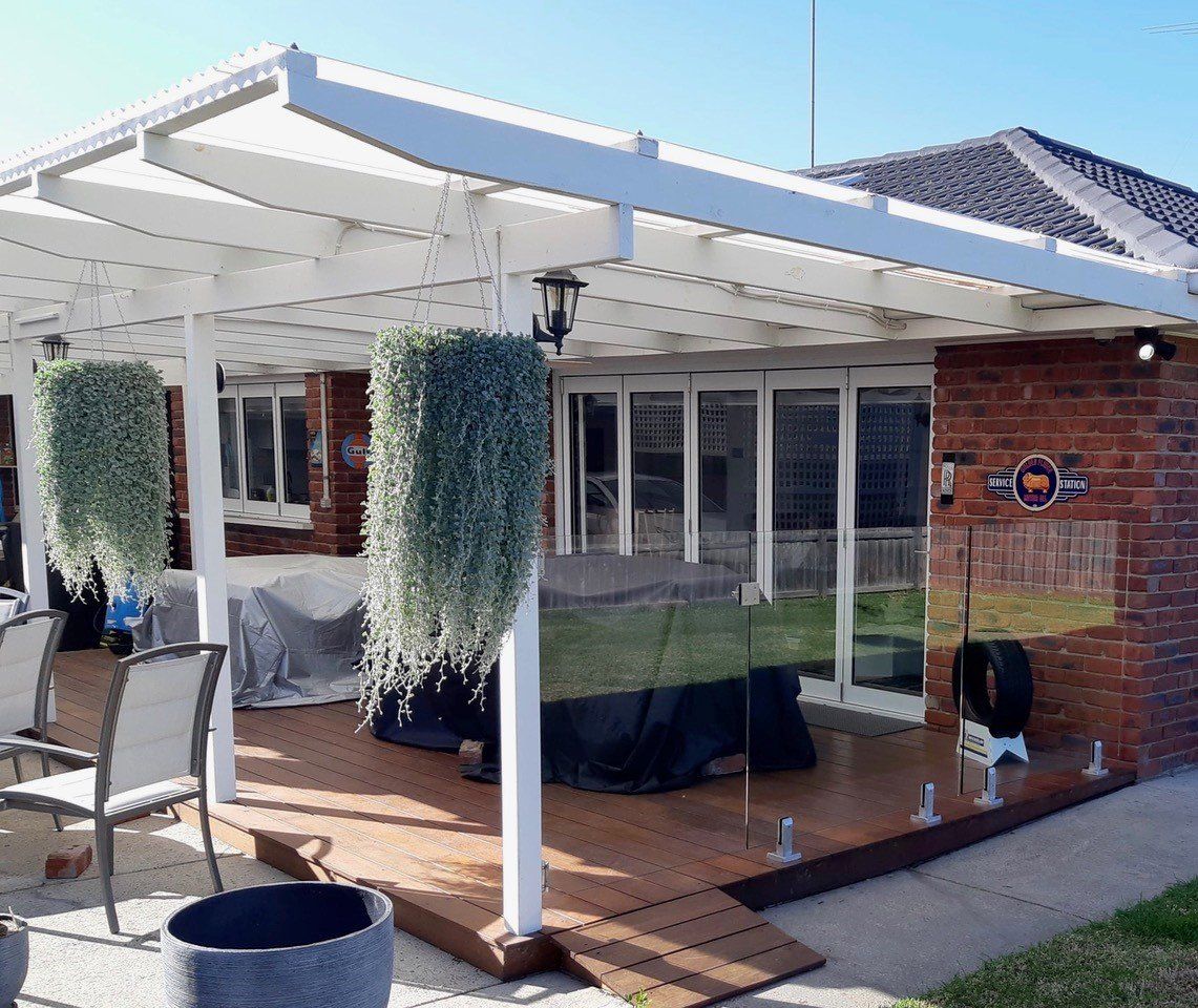 A wooden deck with a white pergola, hanging plants, patio furniture, and a glass fence against a brick house wall.