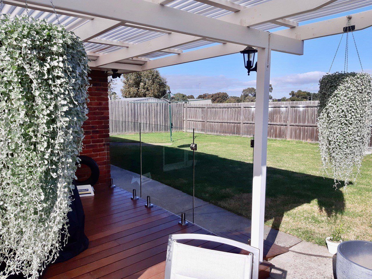 A patio with a wooden deck, a glass balustrade, and hanging plants under a white pergola, overlooking a grassy backyard.