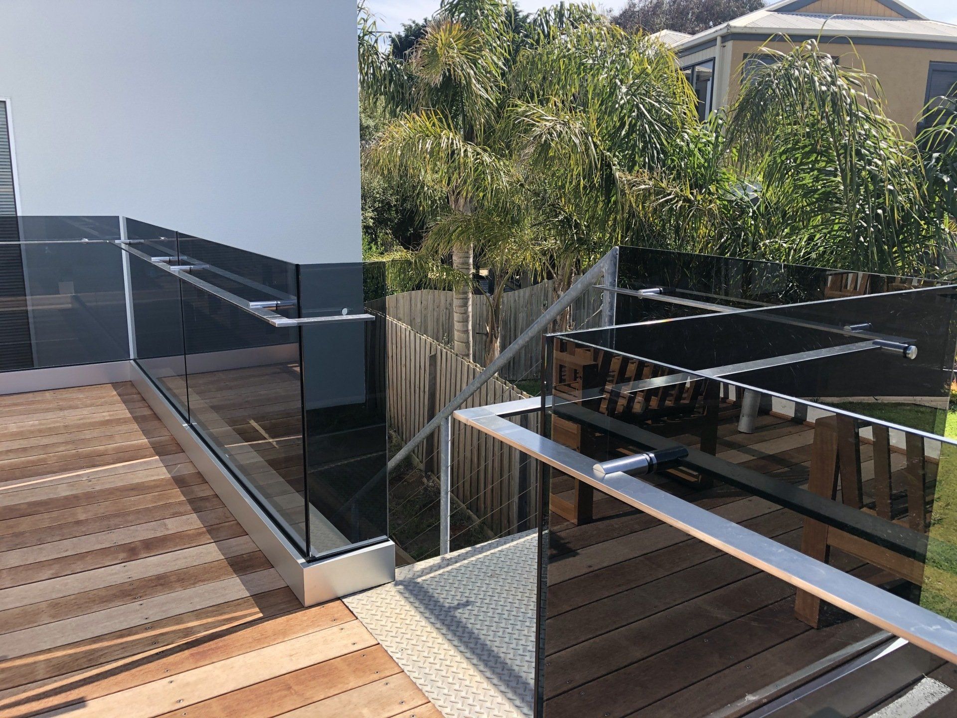 A wooden deck features modern, dark-tinted glass railings with silver metal posts and handrails, overlooking a yard.