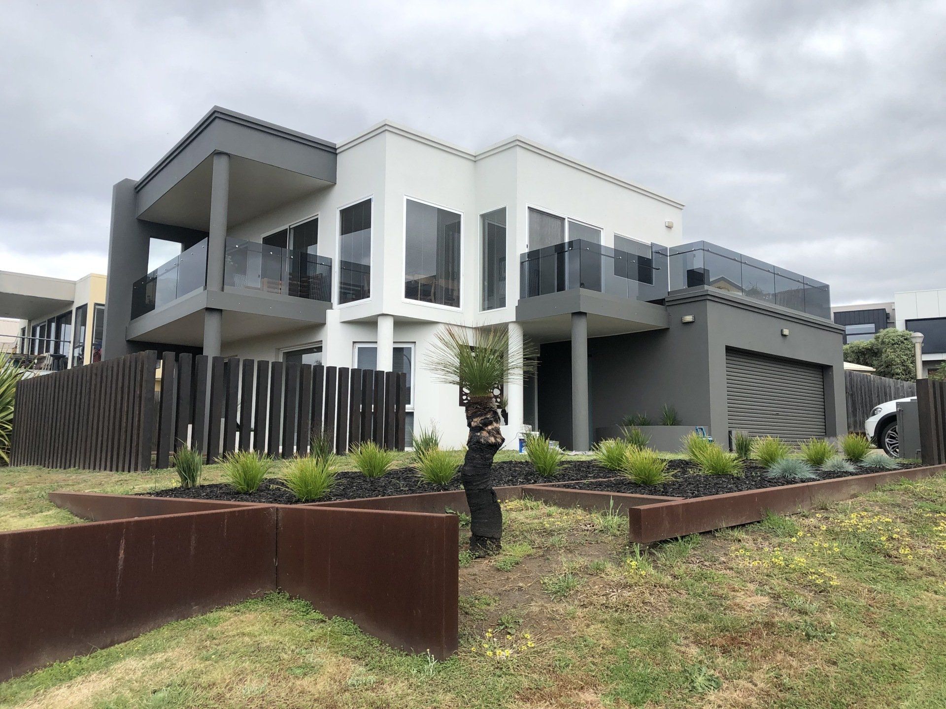 A modern two-story home with white and dark gray exterior, balconies, glass railings, and a landscaped front yard.