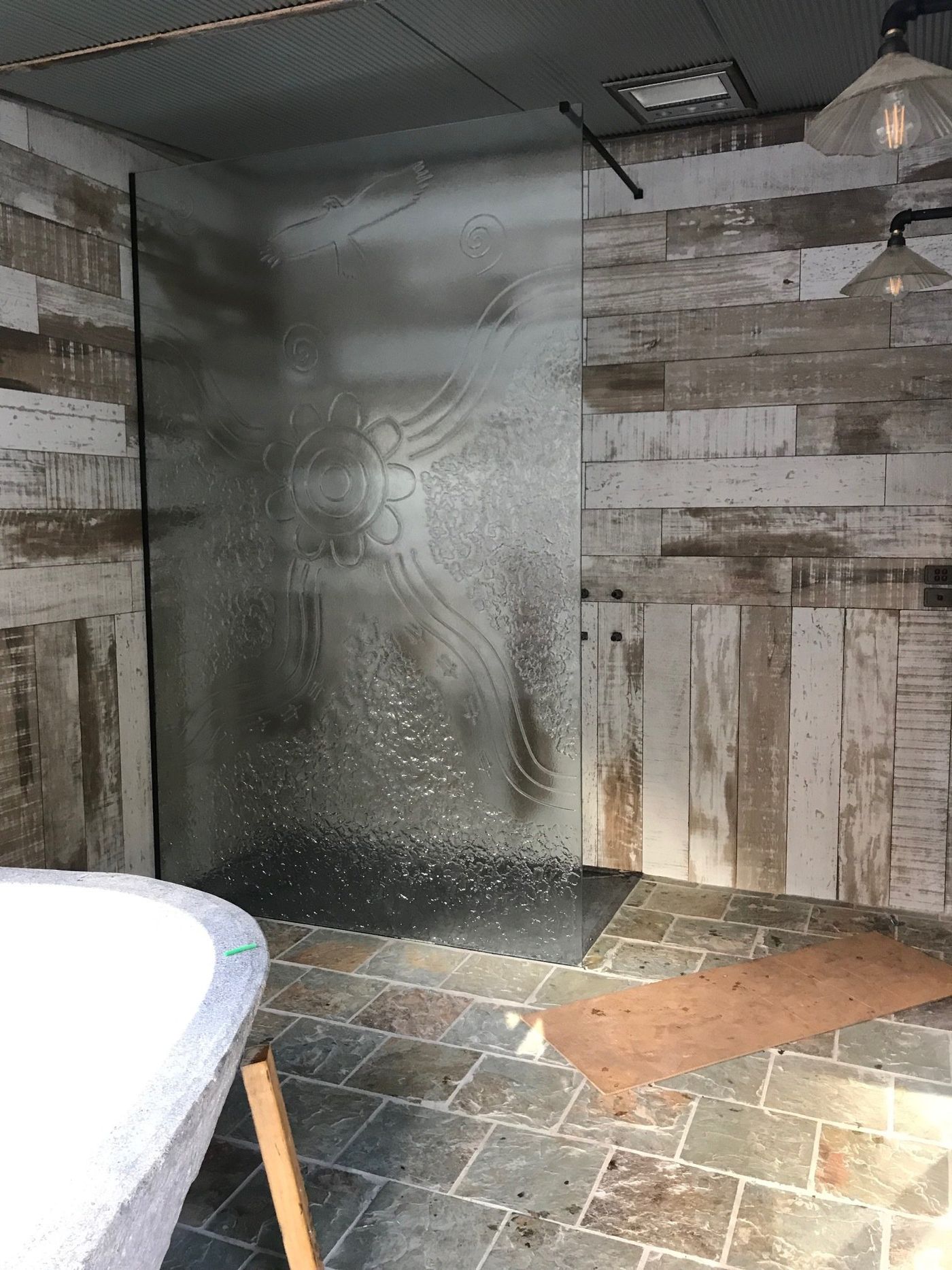 A rustic bathroom with a frosted glass walk-in shower, weathered wood-paneled walls, stone tile floor, and a white tub.