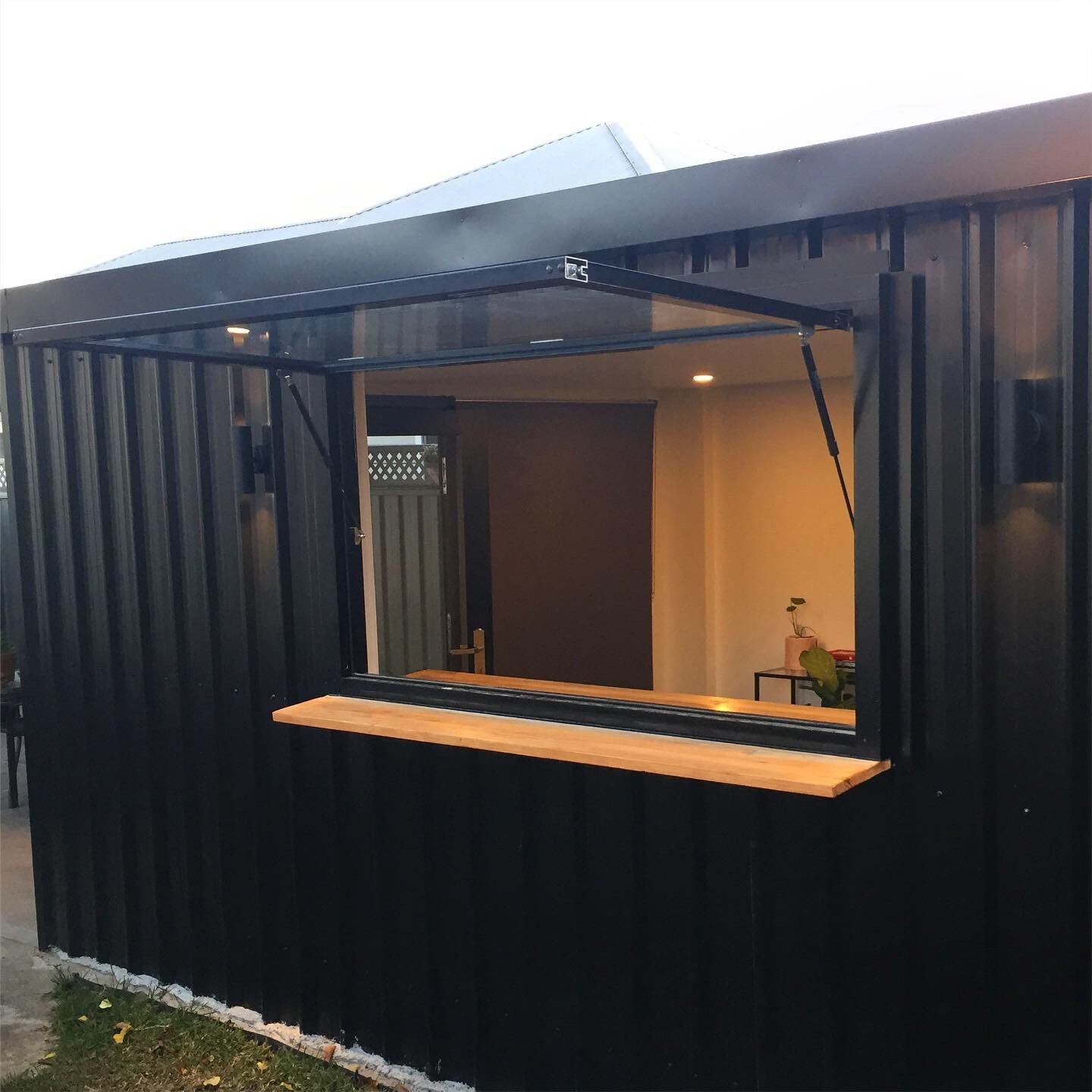 A black, corrugated metal structure featuring an open serving window with a wooden counter and interior lighting.