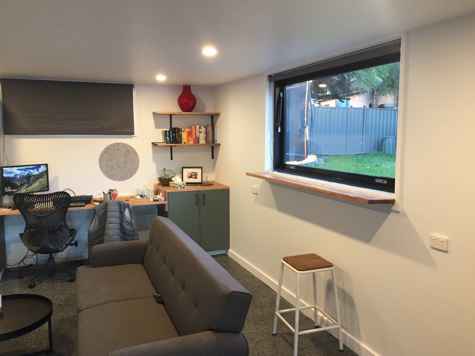 A room featuring a workspace with a computer, a gray couch, a wooden shelf under a window, and a tall stool.