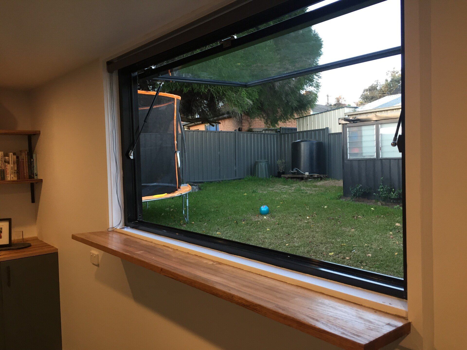 An open window with a wooden sill overlooks a backyard containing a trampoline and a shed.