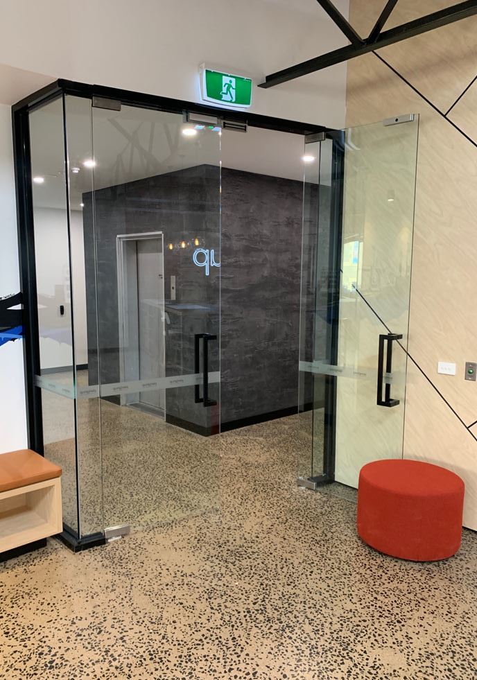 Glass office doors with black frames and handles, leading to an elevator lobby with a dark textured feature wall.