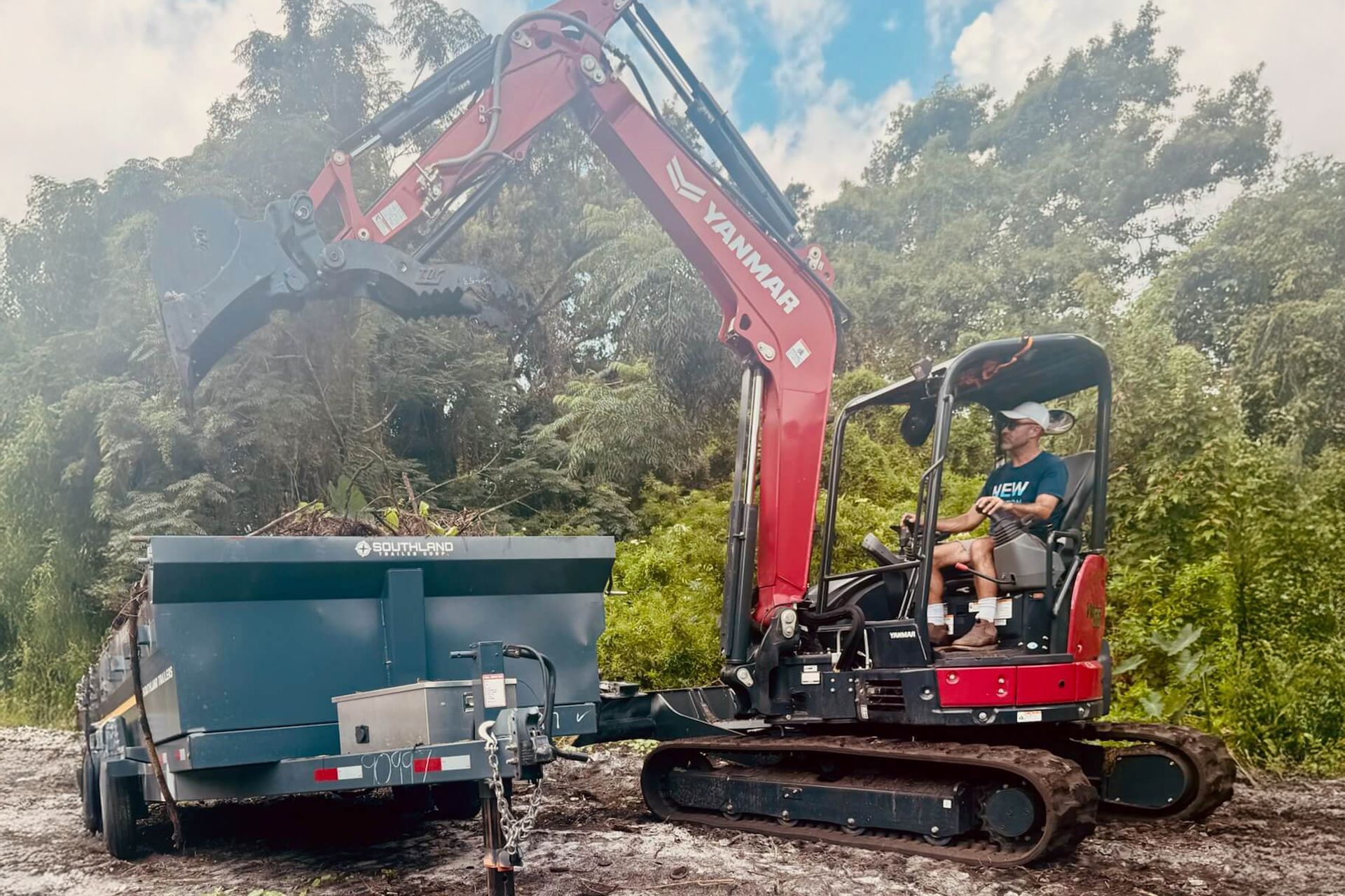 Excavator loading debris into a blue dumpster in a wooded worksite