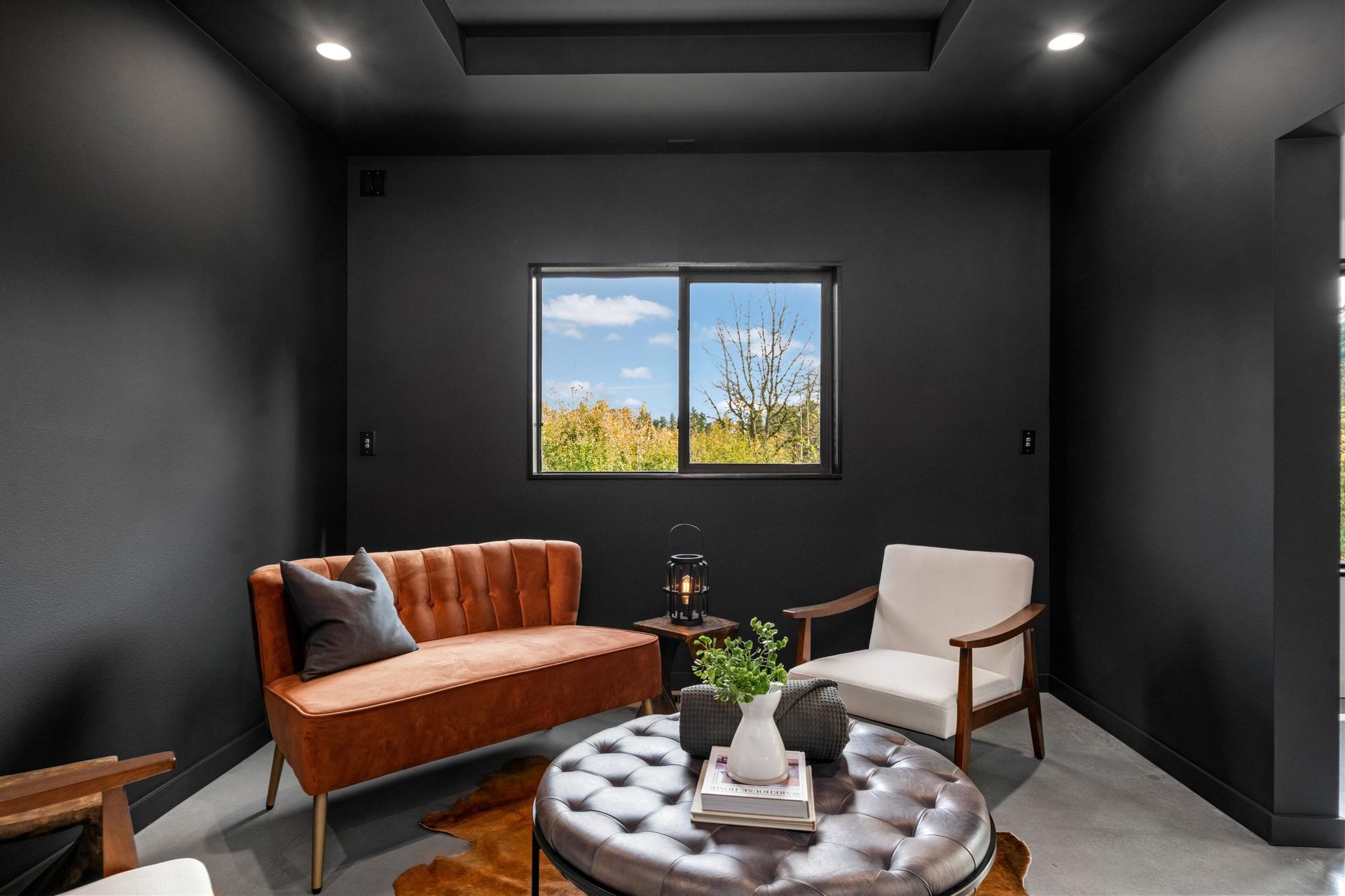 Modern black living room with rust sofa, beige chair, round tufted ottoman, and window view.