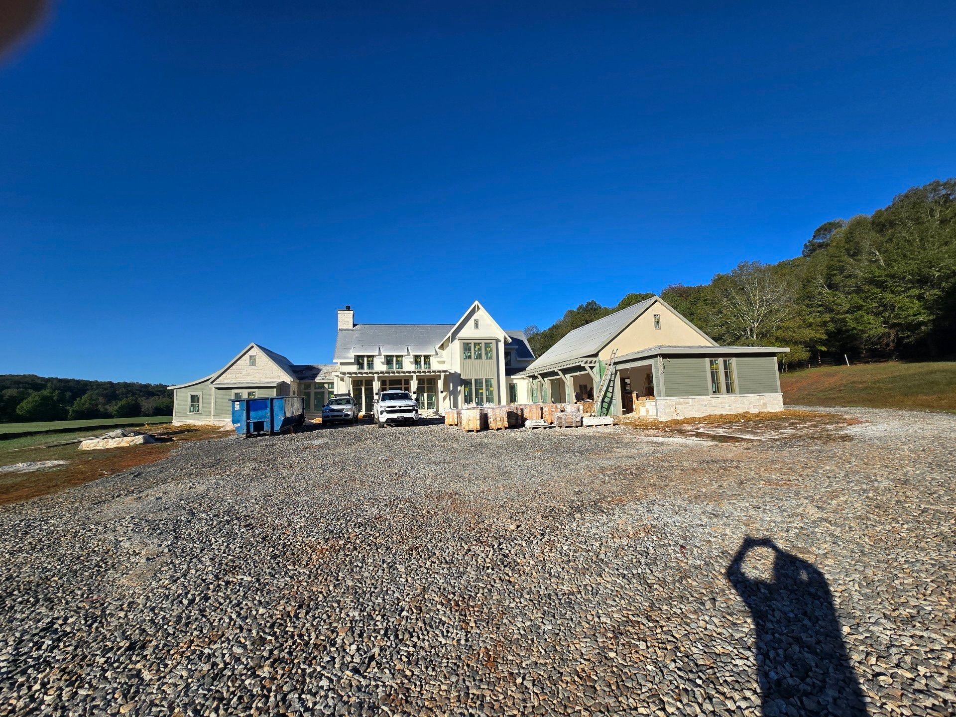 New house under construction, light-colored siding, gravel driveway, blue sky, surrounded by trees and field.