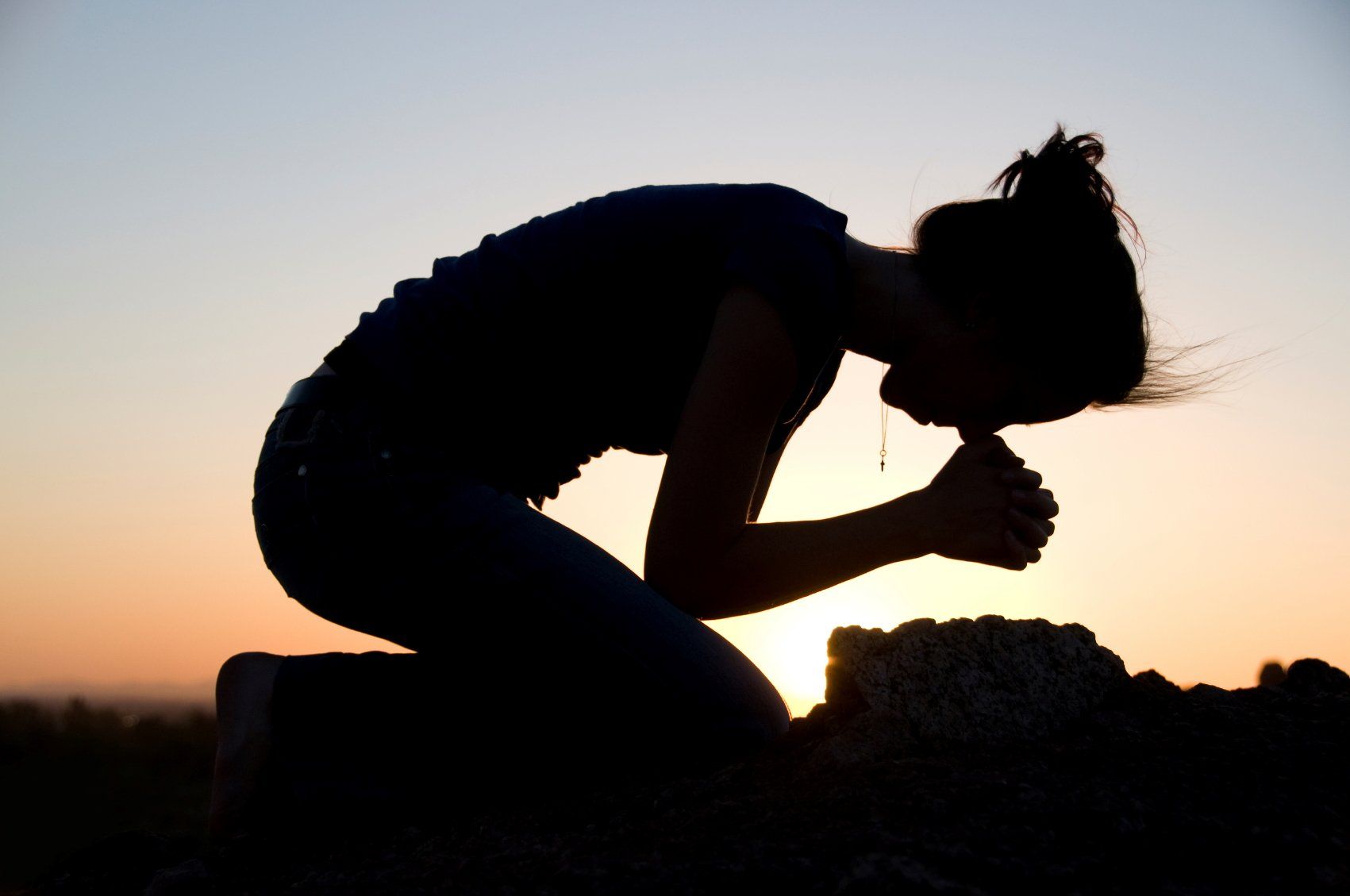Silhouette of person kneeling, head bowed, hands clasped, facing sunset.