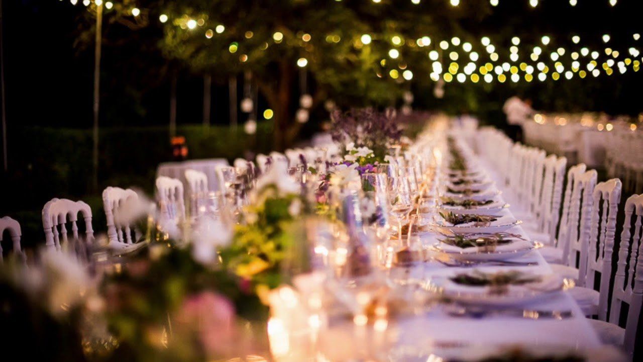 Long tables set for a dinner party, decorated with flowers and fairy lights, outdoors at night.