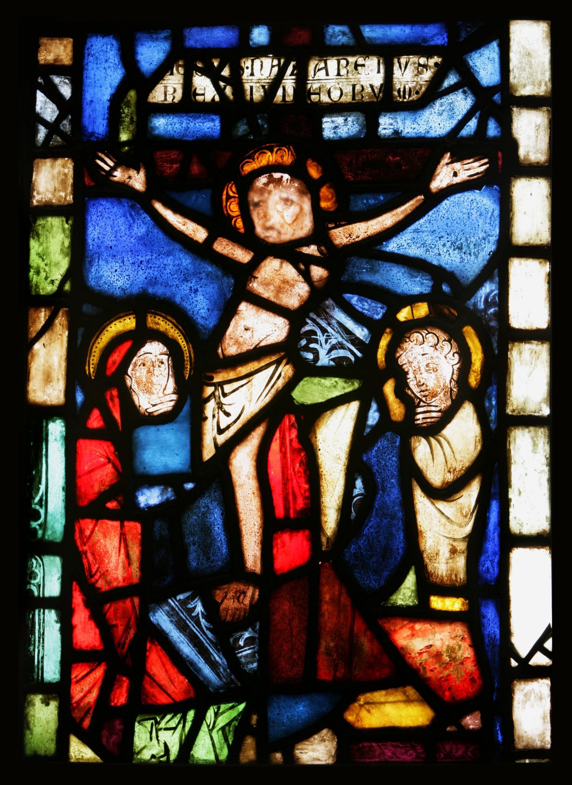 Stained glass depicting Christ on the cross, flanked by two figures, set against a blue and colorful background.
