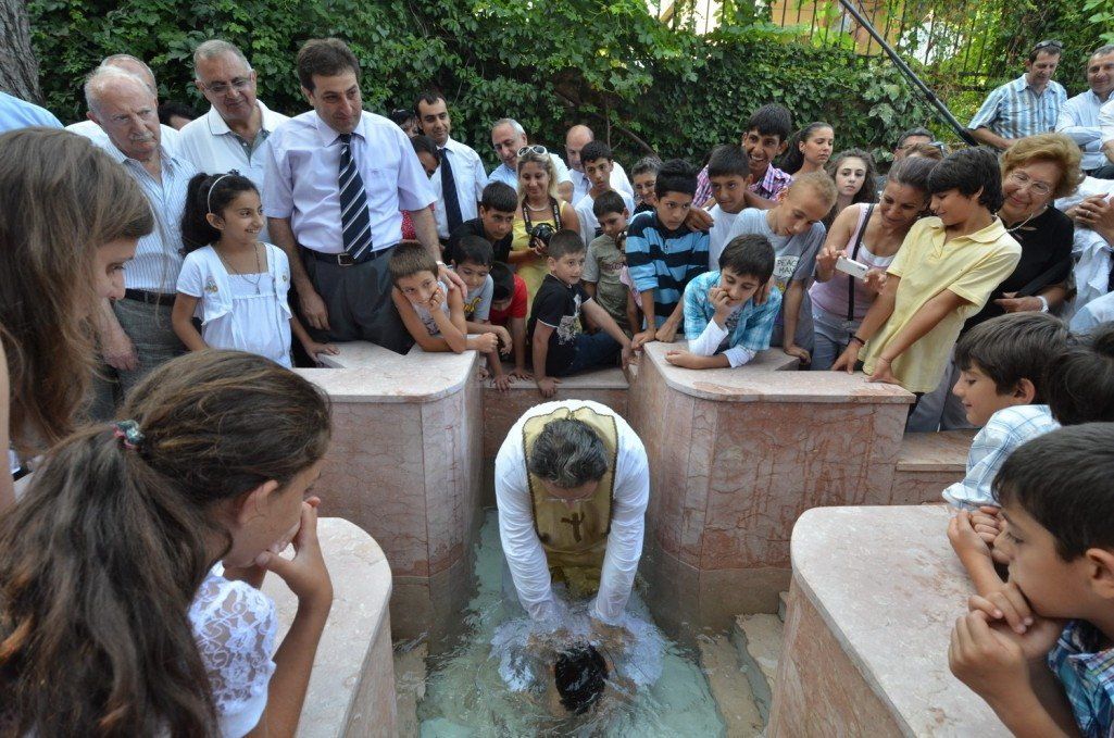 People watching a baptism in an outdoor pool. A person is submerged, wearing a white robe.