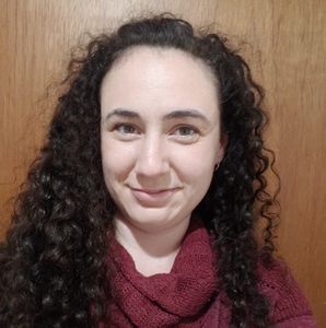 Woman with curly dark hair smiles, wearing a burgundy scarf, against a wood-panel wall.