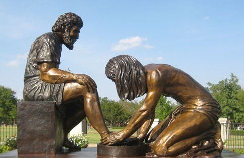 Bronze statue of a person kneeling and washing the feet of another seated person. Outdoor setting.