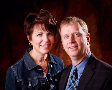 Woman in denim jacket and man in suit smile together against a dark background.