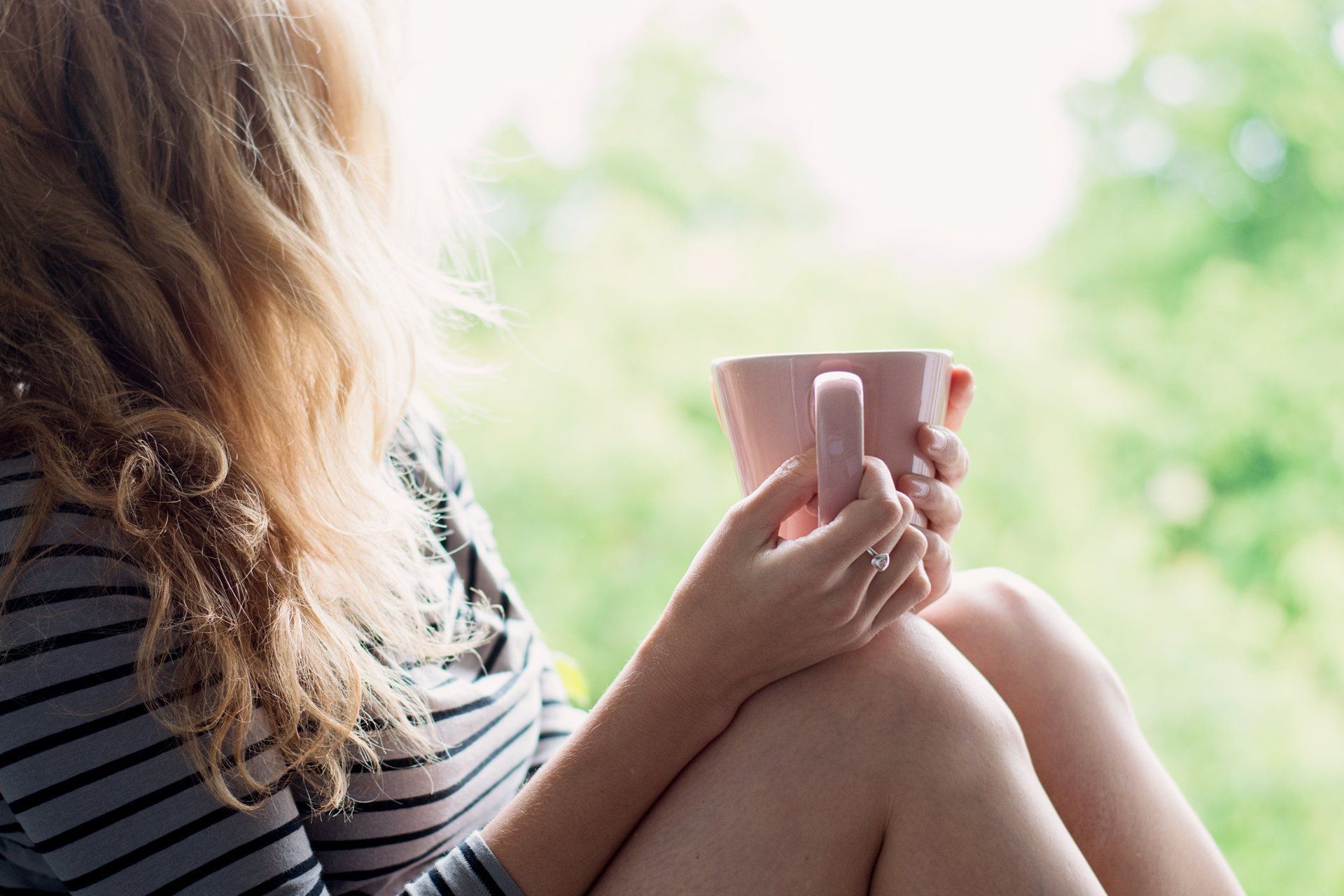 Woman holding a pink mug, looking out a window with green foliage in background.