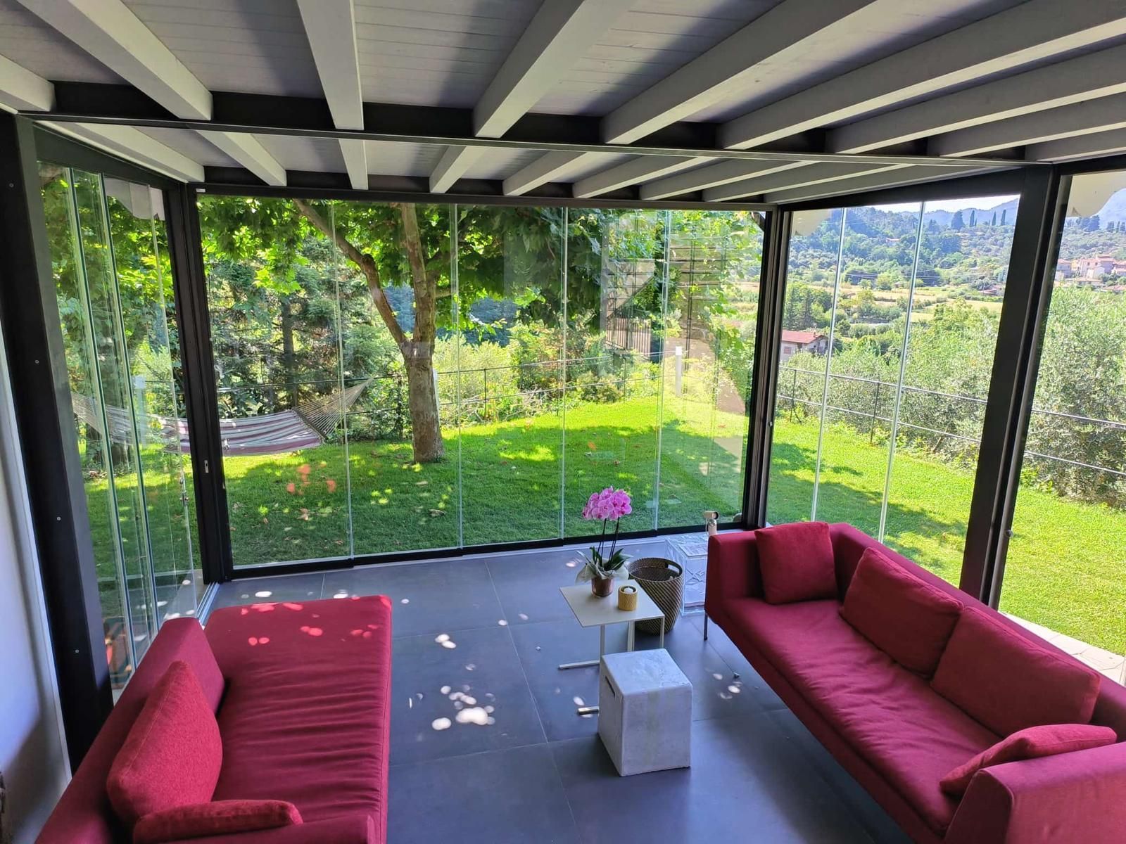 Veranda con pareti in vetro, divani rossi, un tavolino, con vista sul giardino verde e sulla valle.