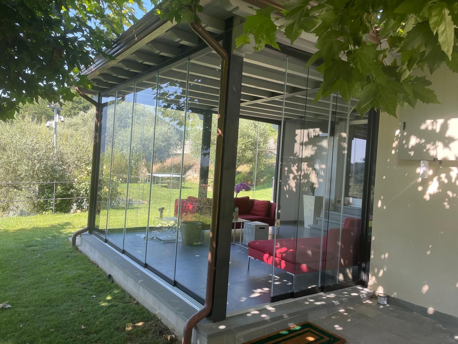 Patio con pareti in vetro, mobili rossi all'interno, circondato da erba verde e alberi.