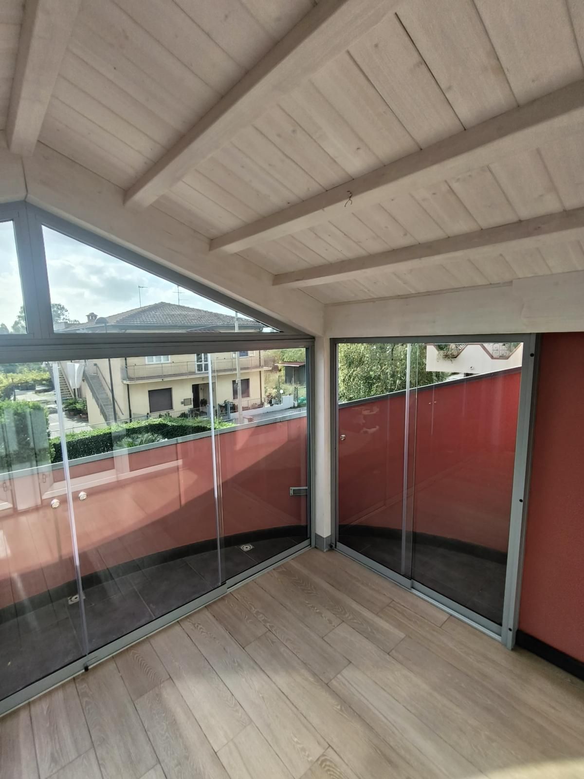 Vista interna di una veranda con ampie vetrate, pavimento e soffitto in legno. Pareti rosse.