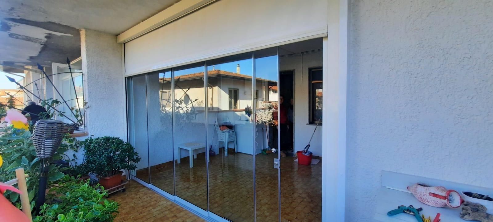 Balcone con vetrate e riflessi, piante sulla sinistra e oggetti all'interno. Giornata di sole.