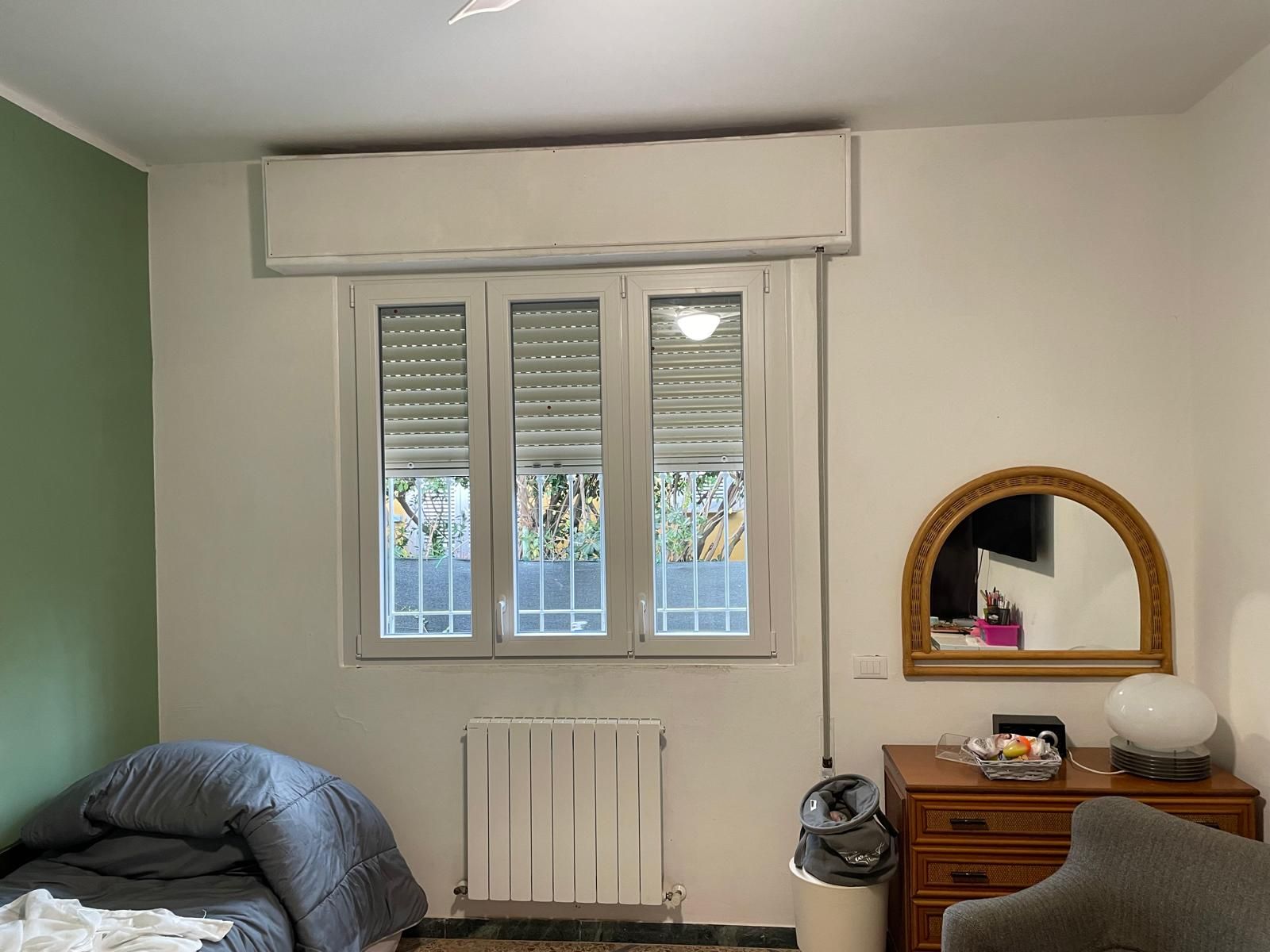 Camera da letto con finestra, aria condizionata, letto, termosifone, cassettiera, specchio e cestino.