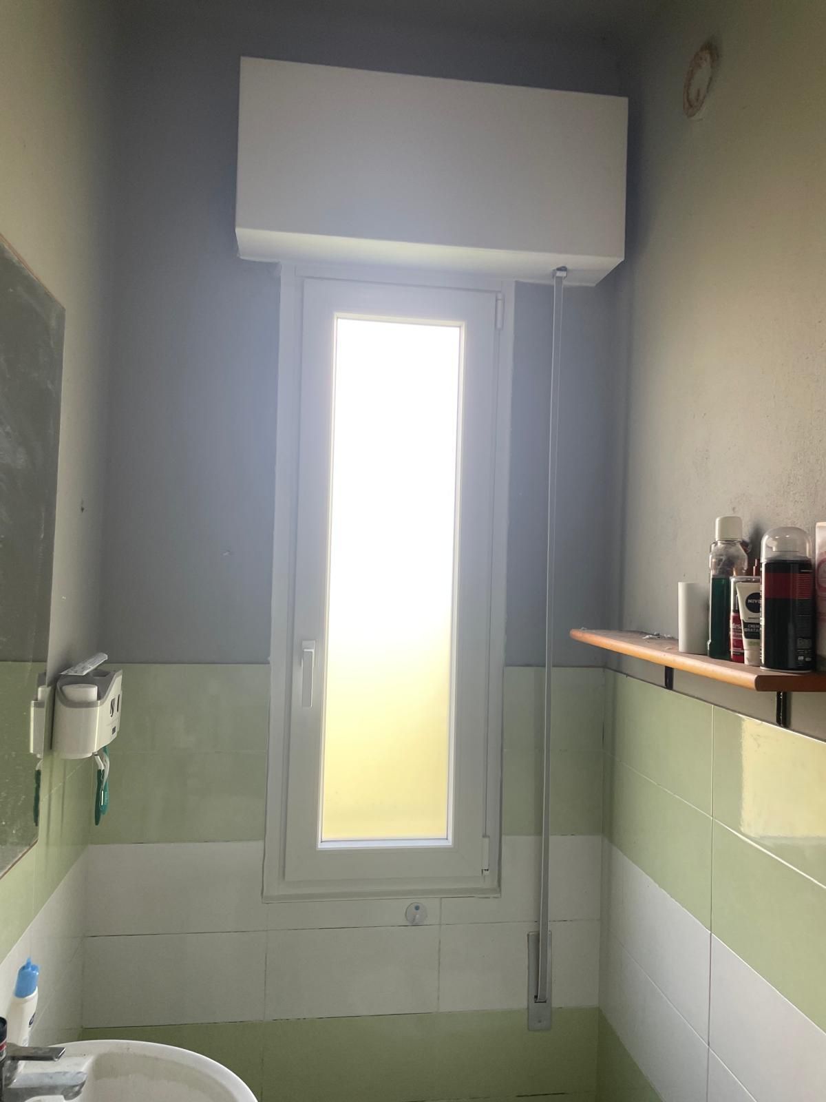 Un piccolo bagno con una finestra alta. Aria condizionata sopra la finestra. Pareti piastrellate di verde, grigio e bianco.