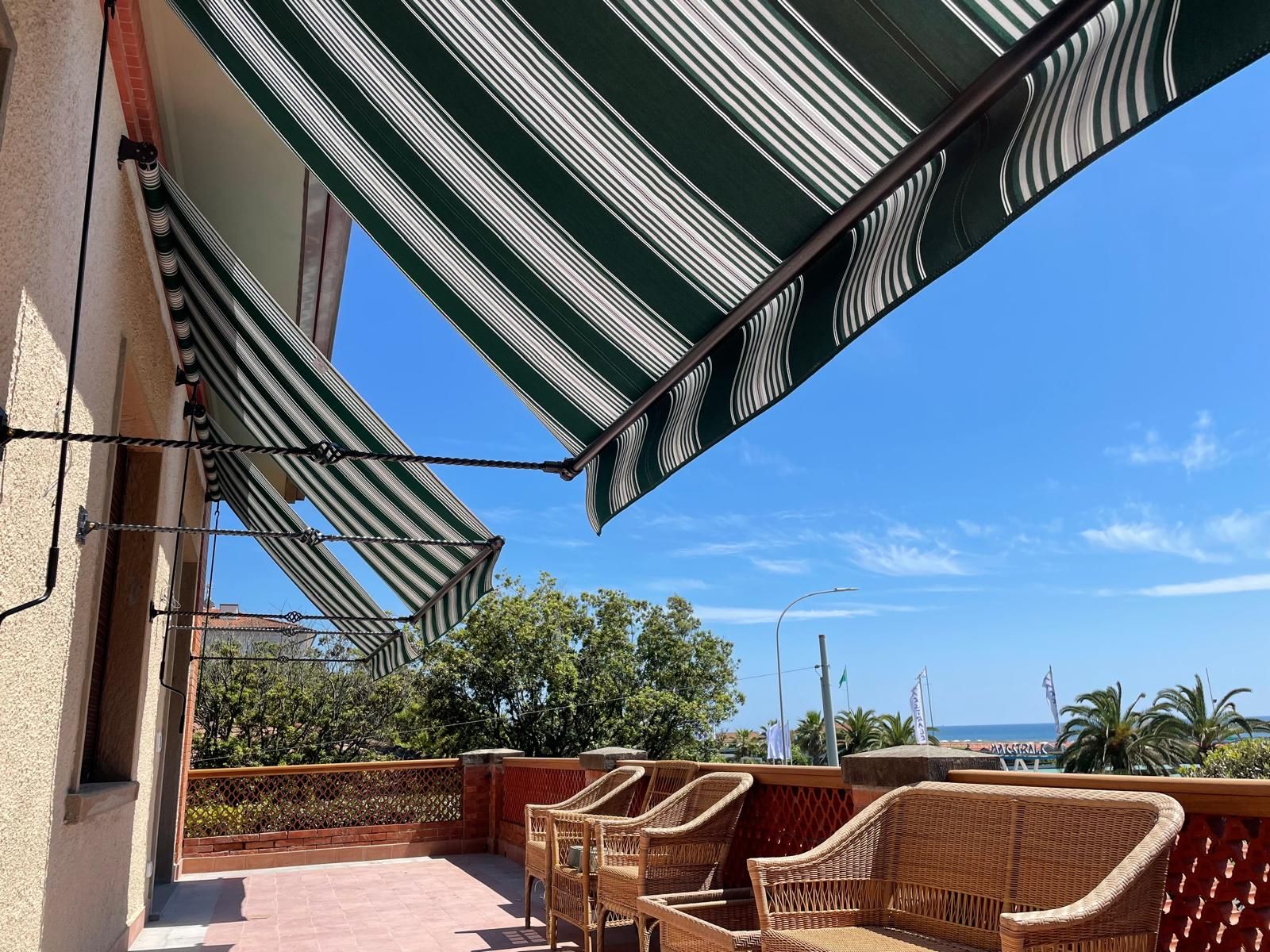 Tende a strisce verdi e bianche ombreggiano un patio con sedie in vimini e vista sull'oceano.