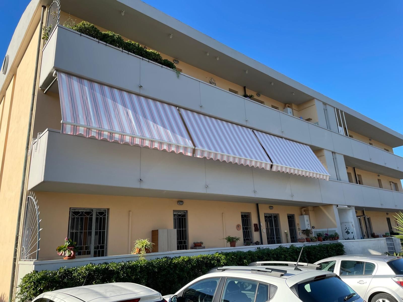 Condominio con tende da sole. Balconi con tende a strisce; auto parcheggiate e arbusti sotto.