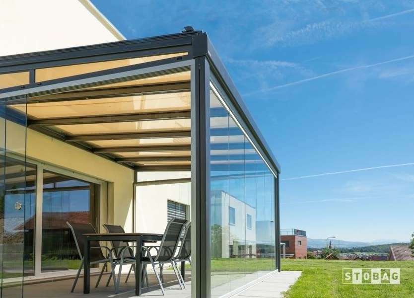 Patio con pareti in vetro, tavolo e sedie, tetto retrattile color marrone chiaro, con cielo azzurro e prato.