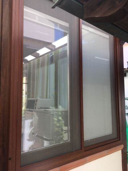 Finestra con cornice marrone e persiana parziale. Il vetro riflettente mostra un interno con tende e mobili.