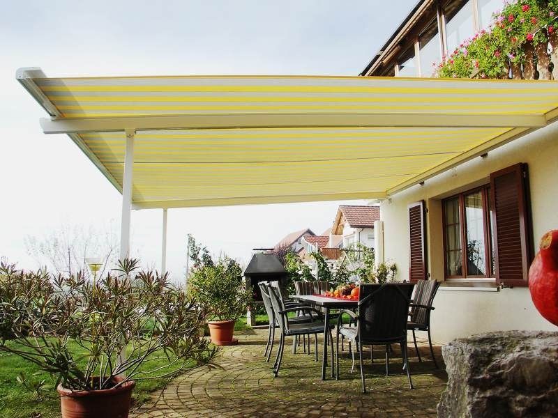 Patio coperto con tavolo e sedie, con vista sul giardino. Tenda da sole gialla, persiane marroni, cielo coperto.