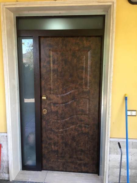 Porta d'ingresso marrone testurizzata con finestra laterale e sopraluce, inserita in un telaio bianco su una parete gialla.